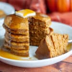 Feature image for our pumpkin panckes show a stack of pancakes on a serving plate with a pat of butter on top and lots of maple syrup; a slice has been taken out and is sitting on the plate next to the stack.