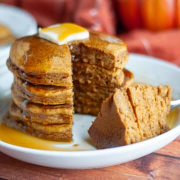 Feature image for our pumpkin panckes show a stack of pancakes on a serving plate with a pat of butter on top and lots of maple syrup; a slice has been taken out and is sitting on the plate next to the stack.