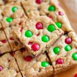 Feature image for our easy christmas cookie bars recipe showing m&m cookie bars cut into squares on a cutting board with a close up on one square stacked on top of the rest.
