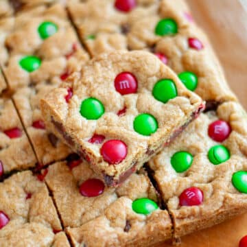 Feature image for our easy christmas cookie bars recipe showing m&m cookie bars cut into squares on a cutting board with a close up on one square stacked on top of the rest.