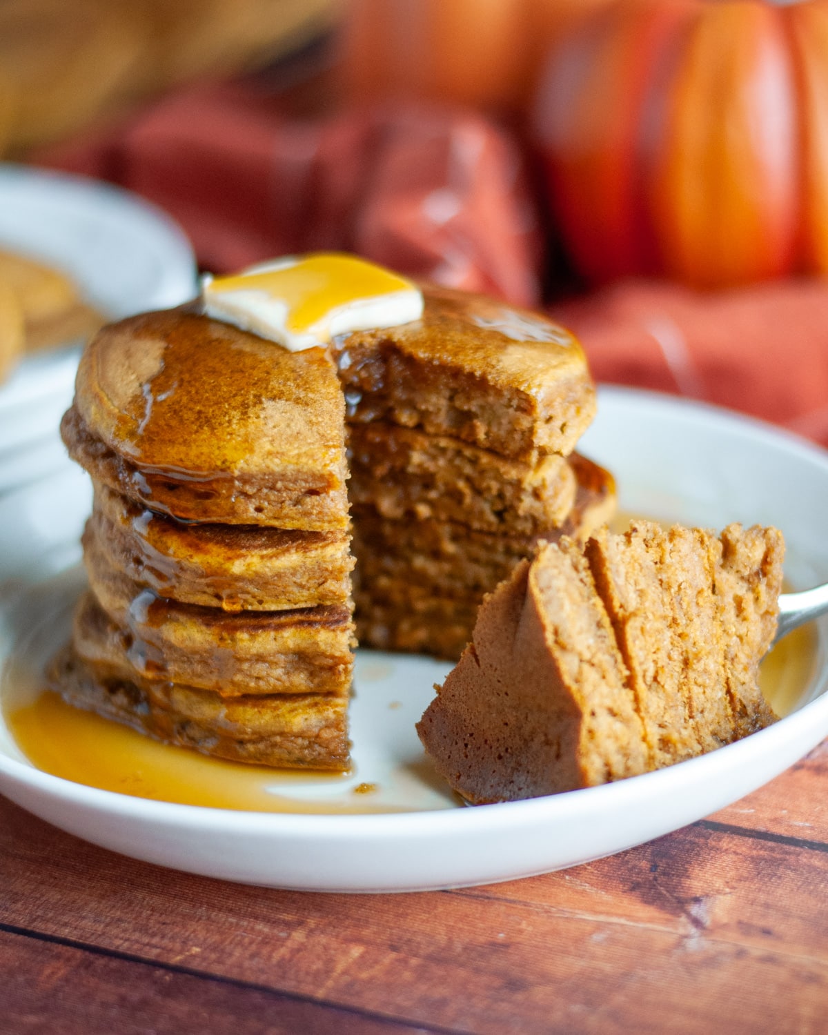 A stack of 4 pancakes on a serving plate with a pat of butter on top and lots of maple syrup; a slice has been taken out and is sitting on the plate next to the stack.