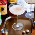 A coupe glass filled with the shaken brandy alexander and topped with freshly grated nutmeg, sitting on a tray surrounded by brandy alexander cocktail ingredients.