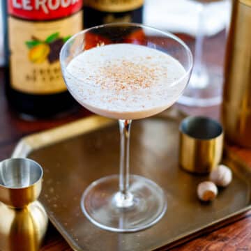 Feature image showing a coupe glass filled with the shaken brandy alexander and topped with freshly grated nutmeg, sitting on a tray surrounded by brandy alexander cocktail ingredients.
