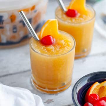 Feature image for our brandy slush drink recipe showing two low ball glasses filled with brandy slushies with a metal straw and garnished with an orange slice and maraschino cherry.
