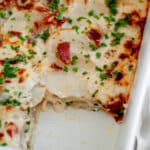 Feature image for our loaded scalloped potatoes, showing the pan of potatoes with a slice taken out of it to showcase the layers of potatoes with cheese dripping out.