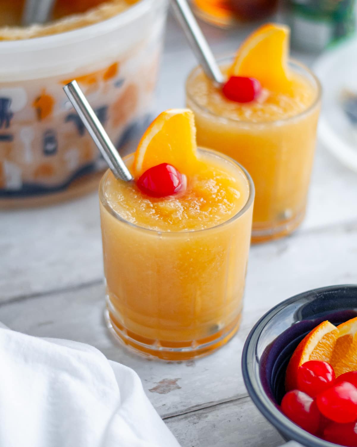 Two low ball glasses filled with Wisconsin brandy slush with a metal straw and garnished with an orange slice and maraschino cherry.