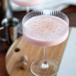 A vibrant pink drink in a coupe glass topped with freshly grated nutmeg, with an additional drink and ingredients around it..