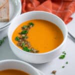 Close up of a bowl of roasted butternut squash soup with carrot topped with pepitas and fresh parsley, with a plate of bread in the background.