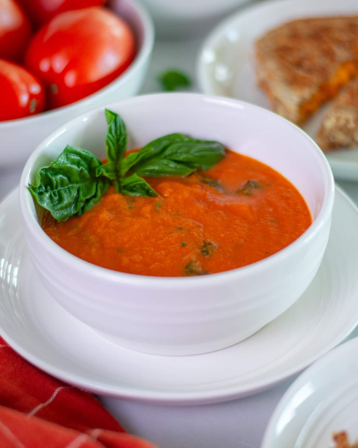 A bowl of freshly made tomato & basil soup with a sprig of basil for garnish, with extra roma tomatoes, and a grilled cheese sandwich in the background.