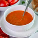A bowl of freshly made tomato basil soup with a spoon in it, with extra roma tomatoes, and a grilled cheese sandwich in the background.