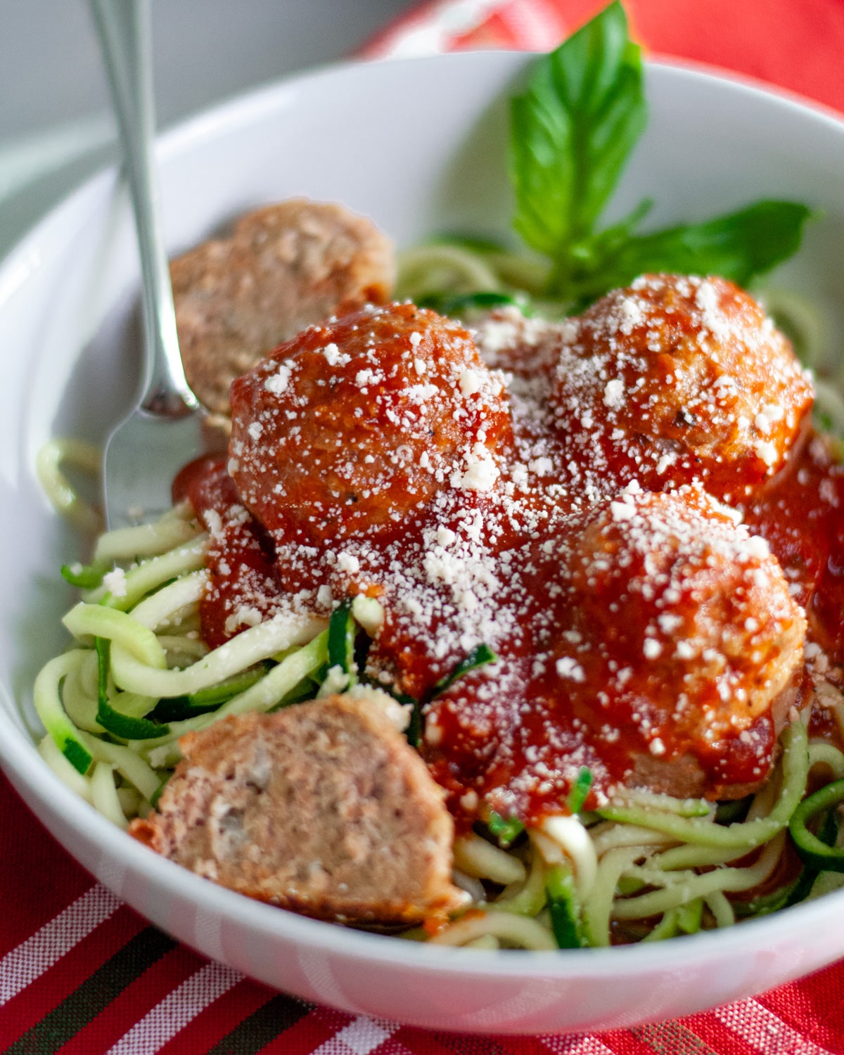 A white dish filled with zucchini noodles topped with red sauce, ground turkey meatballs, and grated parmesan cheese with a fork twirled into the noodles and a fresh basil garnish.