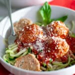 A white dish filled with zucchini noodles topped with red sauce, ground turkey meatballs, and grated parmesan cheese with a fork twirled into the noodles and a fresh basil garnish.