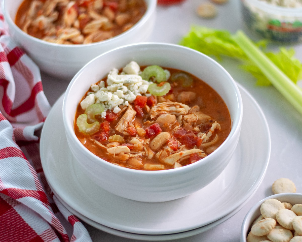 A bowl of white chicken chili topped with blue cheese and some celery pieces, surrounded by a red and white linen, celery stalks, oyster crackers, and a carton of blue cheese crumbles.