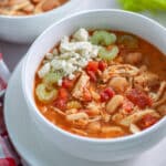 A bowl of white chicken chili topped with blue cheese and some celery pieces, surrounded by a red and white linen, and celery stalks.