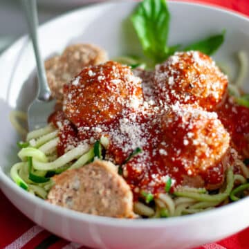A white dish filled with zucchini noodles topped with red sauce, ground turkey meatballs, and grated parmesan cheese with a fork twirled into the noodles and a fresh basil garnish.