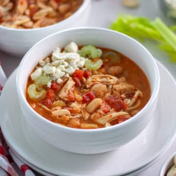 A bowl of white chicken chili topped with blue cheese and some celery pieces, surrounded by a red and white linen, celery stalks, and oyster crackers.