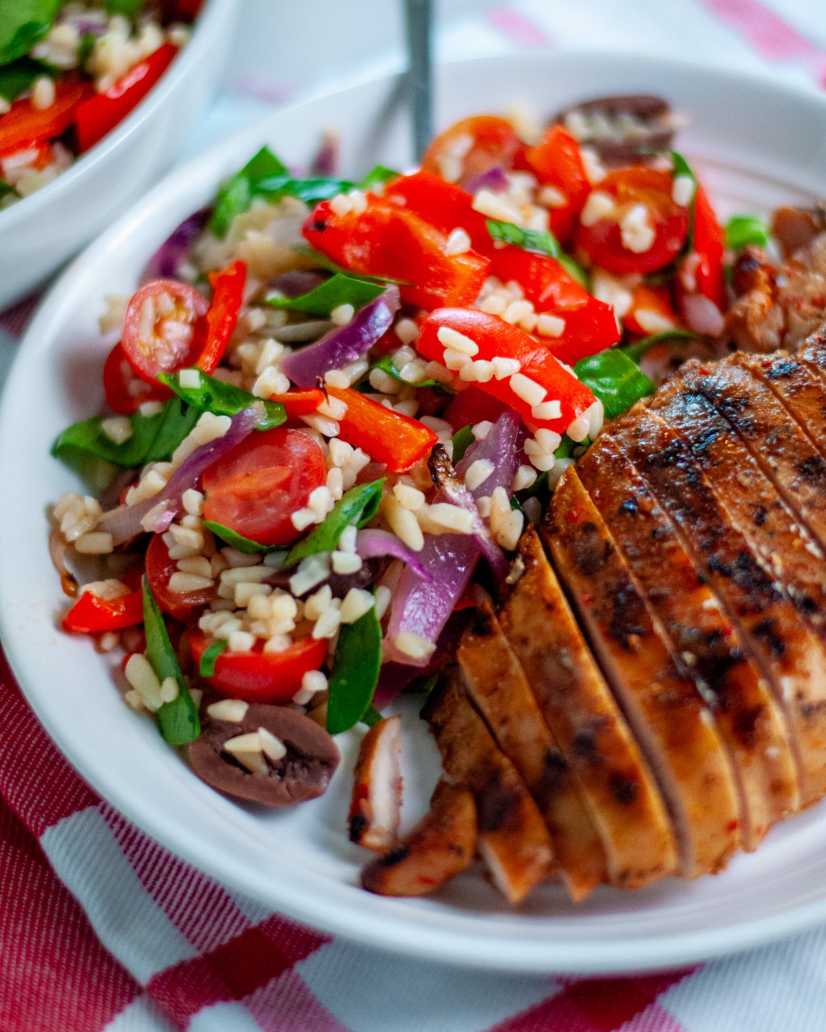 A serving plate filled with mediterranean pasta salad with orzo and a sliced, grilled chicken breast.