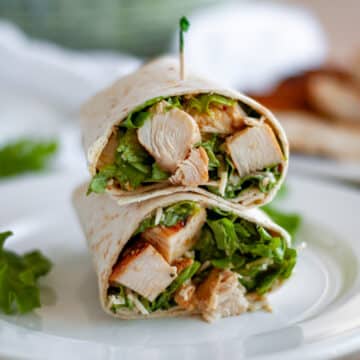 A chicken caesar salad wrap cut in half and stacked on top of each other on a white serving plate with a toothpick stuck into the top wrap.