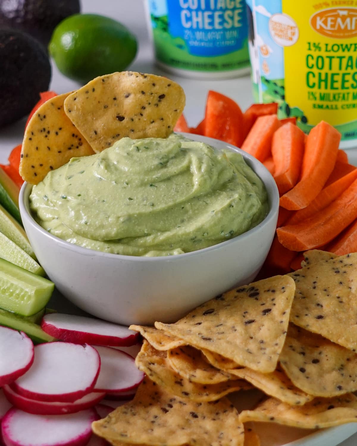 Close up of a bowl of high protein avocado dip on a veggie tray with two tortilla chips dunked into the dip.