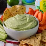 Close up of a bowl of high protein avocado dip on a veggie tray with two tortilla chips dunked into the dip.