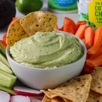 Close up of a bowl of high protein avocado dip on a veggie tray with two tortilla chips dunked into the dip.