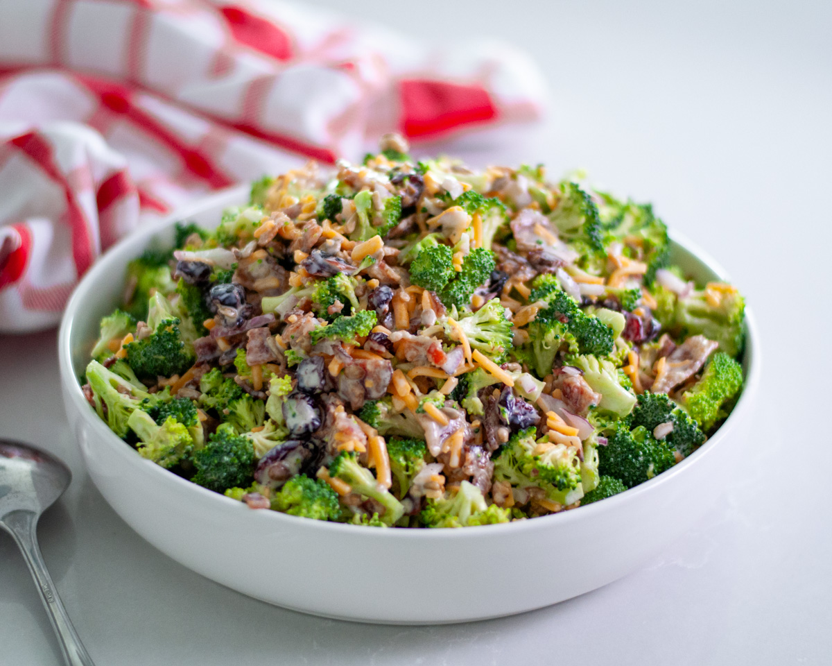 A serving bowl filled high with broccoli salad with bacon with a serving spoon next to it and a checkered linen behind it.