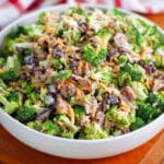A serving bowl filled high with broccoli salad on a wooden cutting board with a checkered linen behind it.