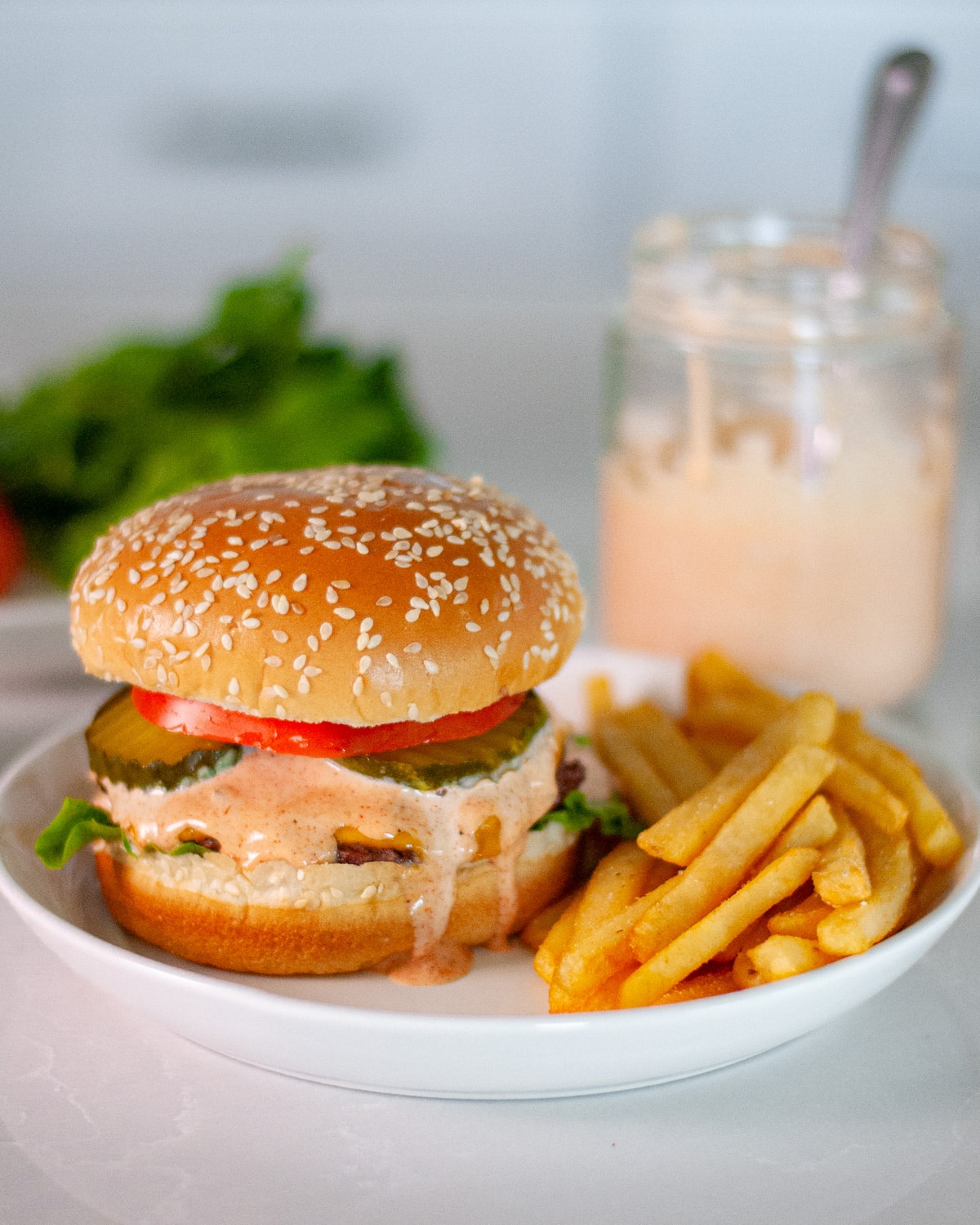 Head-on shot of a cheeseburger dripping with homemade burger sauce, pickles, tomato, and lettuce on a sesame seed bun with fries.