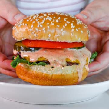 Head-on shot of two hands holding cheeseburger dripping with homemade burger sauce, pickles, tomato, and lettuce on a sesame seed bun.