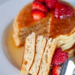 Close up for a stack of 3 pancakes topped with berries and maple syrup with a slice out of it, stacked on a fork to show the interior texture.