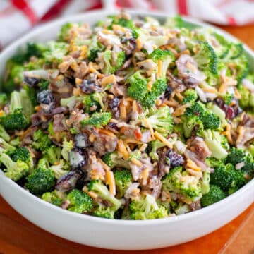 A serving bowl filled high with broccoli salad on a wooden cutting board with a checkered linen behind it.
