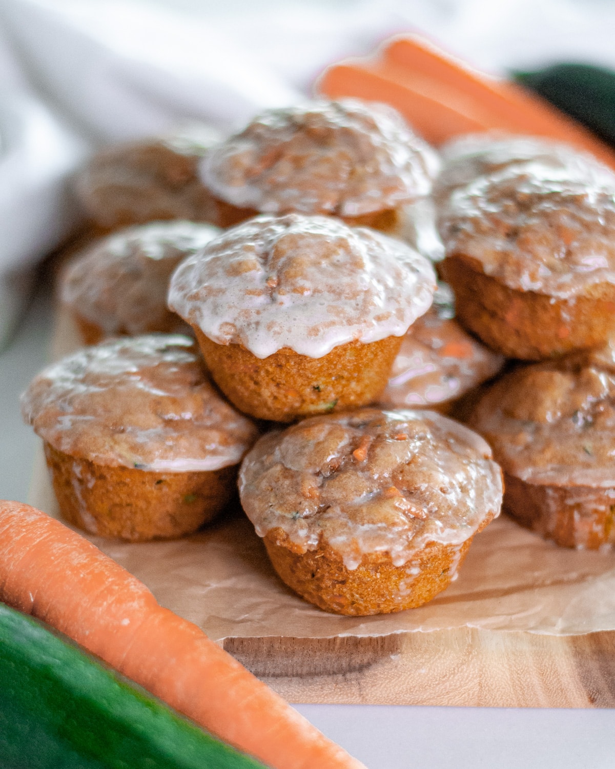 Glazed carrot zucchini muffins stacked on a wooden board, with carrots and zucchinis around it.