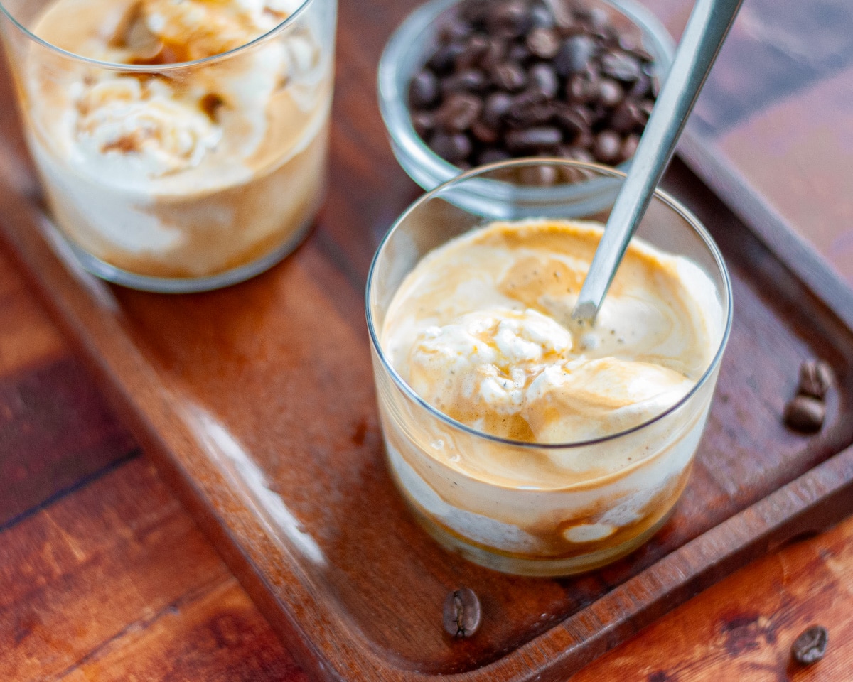 Close up of a freshly made affogato on a wooden tray with espresso beans around it and another affogato in the background.