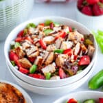 Close up of a bowl of chicken and strawberry salad in a large white serving bowl with salad ingredients around it.
