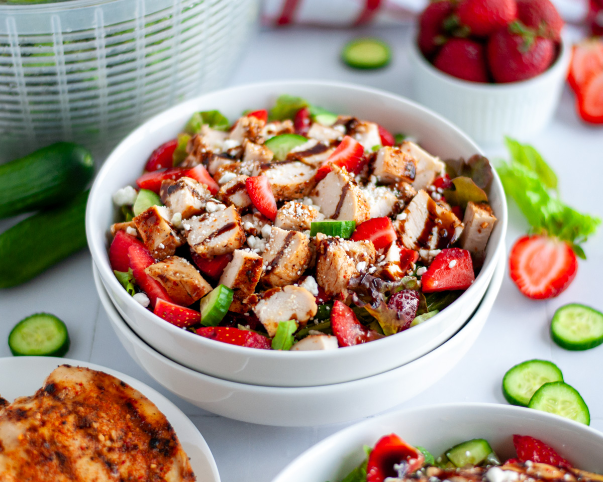 Close up of a bowl of chicken and strawberry salad in a large white serving bowl with salad ingredients around it.