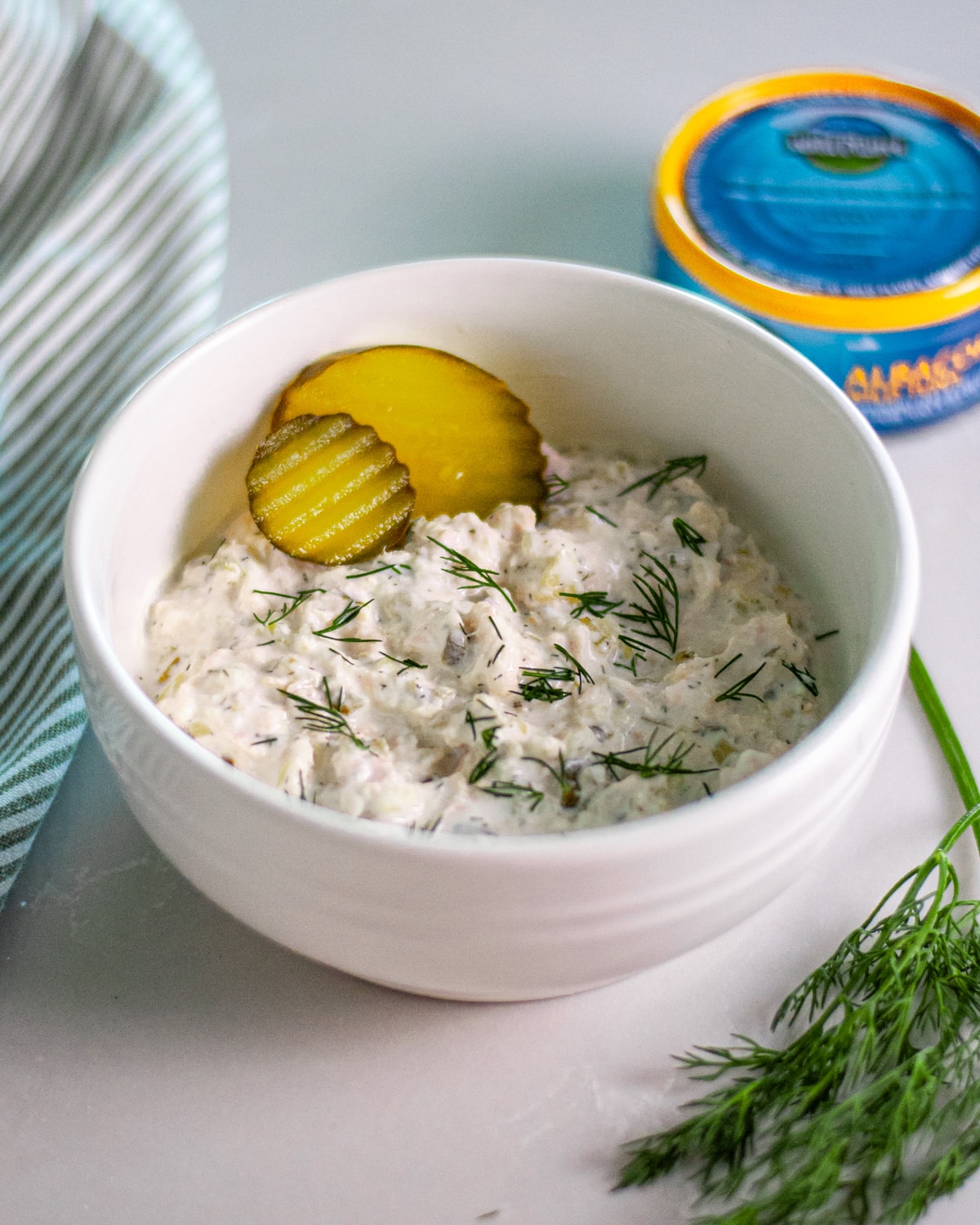 A bowl of creamy tuna salad topped with fresh dill and a couple of pickle rounds with a striped linen, can of tuna, and fresh dill around it.