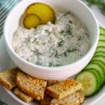A bowl of tuna salad with pickles topped with fresh dill and a couple of pickle rounds on a plate with pieces of toasted bread and cucumber slices.