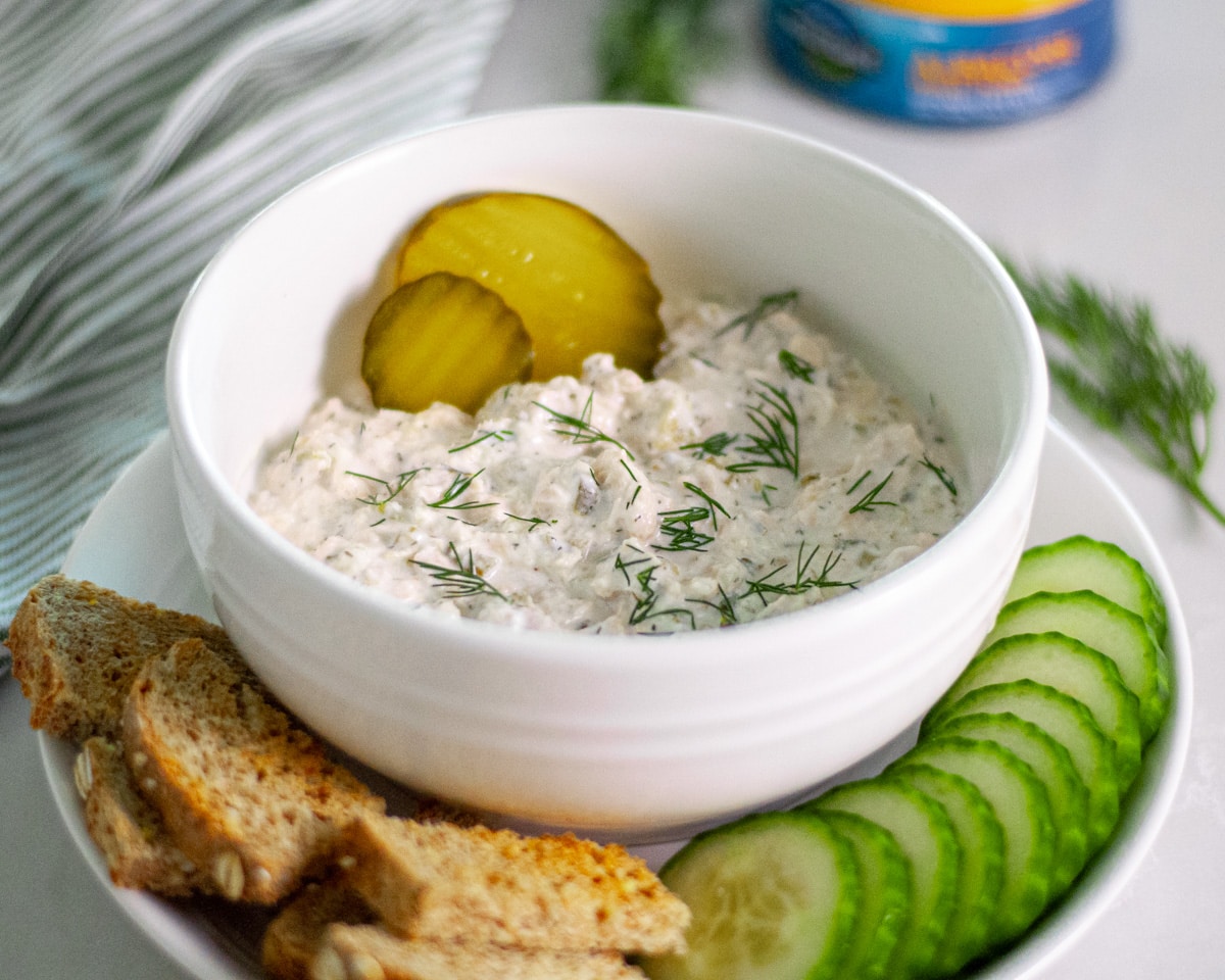 A bowl of tuna salad with pickles topped with fresh dill and a couple of pickle rounds on a plate with pieces of toasted bread and cucumber slices.