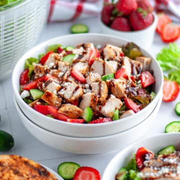 Close up of a bowl of chicken and strawberry salad in a large white serving bowl with salad ingredients around it.
