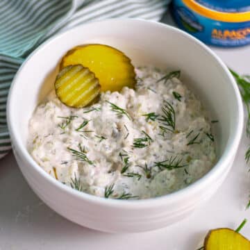 Feature image for our dill pickle tuna salad recipe showing a bowl of creamy tuna salad topped with fresh dill and a couple of pickle rounds with a striped linen, can of tuna, and fresh dill around it.