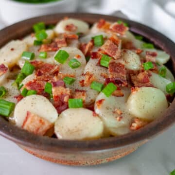A brown pottery serving bowl filled to the brim with hot German potato salad topped with large pieces of bacon and green onion.