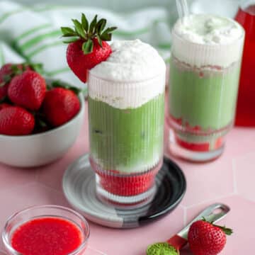 Close up of a freshly made strawberry iced matcha with strawberry cold foam garnished with a fresh strawberry on the rim, surrounded by other ingredients.