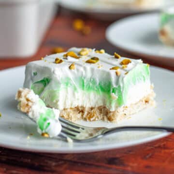 Feature image showing a slice of layered pistachio dessert on a serving plate with a bite taken out of it and a portion resting on a fork on the plate.