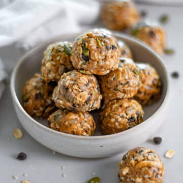Close up of a pile of no bake energy balls with chia seeds, flax, pumpkin seeds, oats, sesame seeds, coconut, sunflower seeds, and peanut butter in a small bowl surrounded by other energy bites, a white linen, and scattered ingredients.