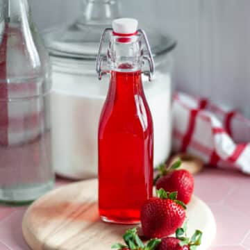 Feature image showing a lidded glass jar filled with strawberry simple syrup, sitting on a wooden board and surrounded by ingredients and a checkered linen.