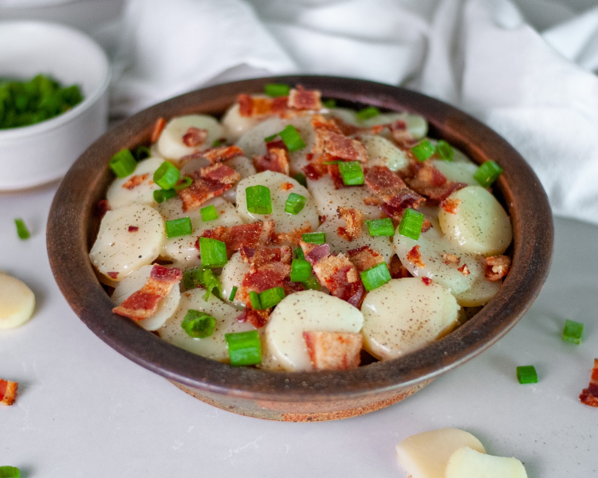 A brown pottery serving bowl filled to the brim with hot German potato salad topped with large pieces of bacon and green onion.