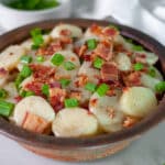 A brown pottery serving bowl filled to the brim with hot German potato salad topped with large pieces of bacon and green onion.