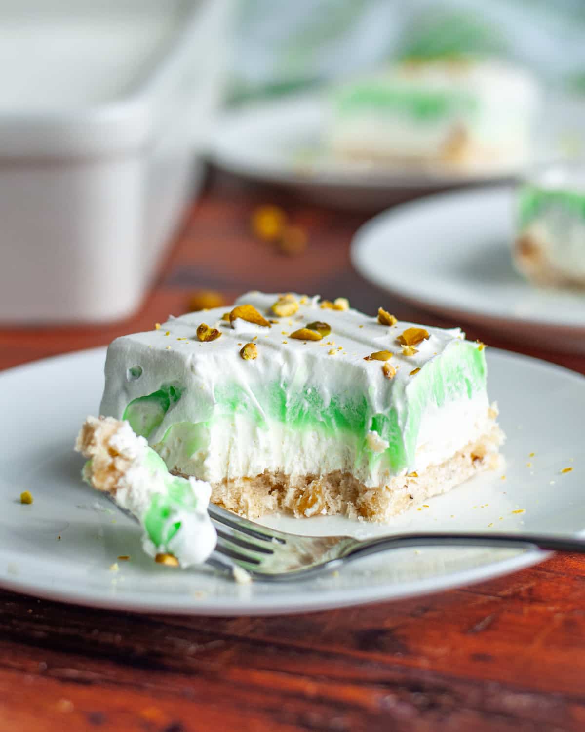 A square of the layered pistachio pudding dessert on a white serving plate, topped with chopped pistachios with a bite of the dessert on a fork resting on the plate.