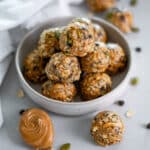 Close up of a pile of no bake energy bites with peanut butter and seeds in a small bowl surrounded by other energy bites, a white linen, a spoon of peanut butter, and scattered ingredients.