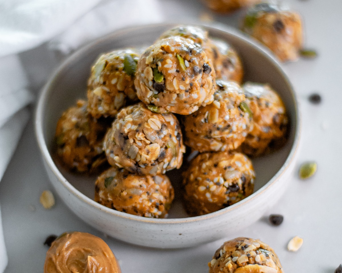Close up of a pile of no bake energy bites with peanut butter and seeds in a small bowl surrounded by other energy bites, a white linen, and scattered ingredients.